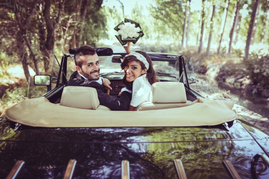 Newlyweds driving away in a convertible car, looking back and smiling.