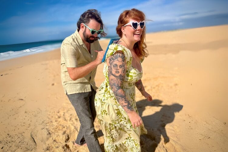 A fun man and woman at the beach wearing love heart sunglasses. The man is using the woman's back to sign the marriage certificate as one of the wedding witnesses
