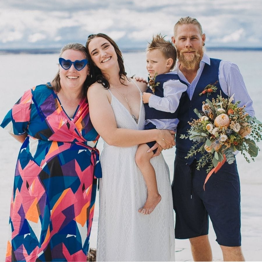 Kate Ryan Taylor with a couple and their child. Kate is wearing a bright patterned dress, love heart glasses, and standing on the beach