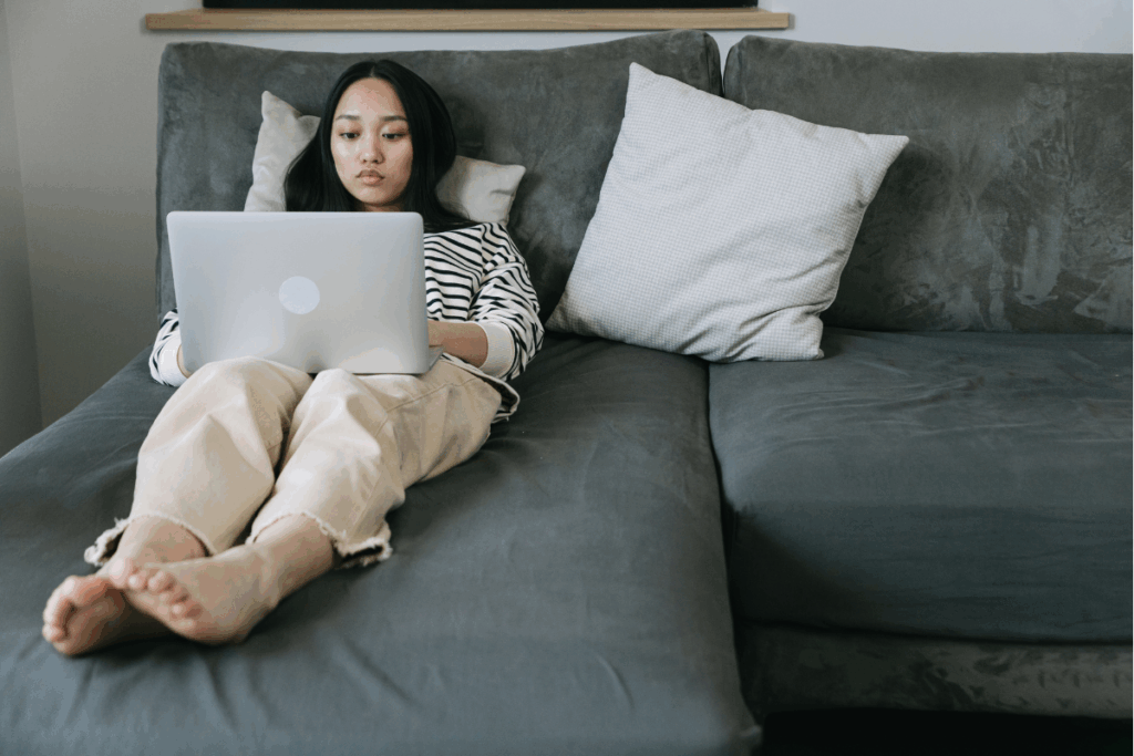 Woman on a dark grey couch in a fully relaxed position looking at her laptop – perhaps she is completing her celebrant OPD