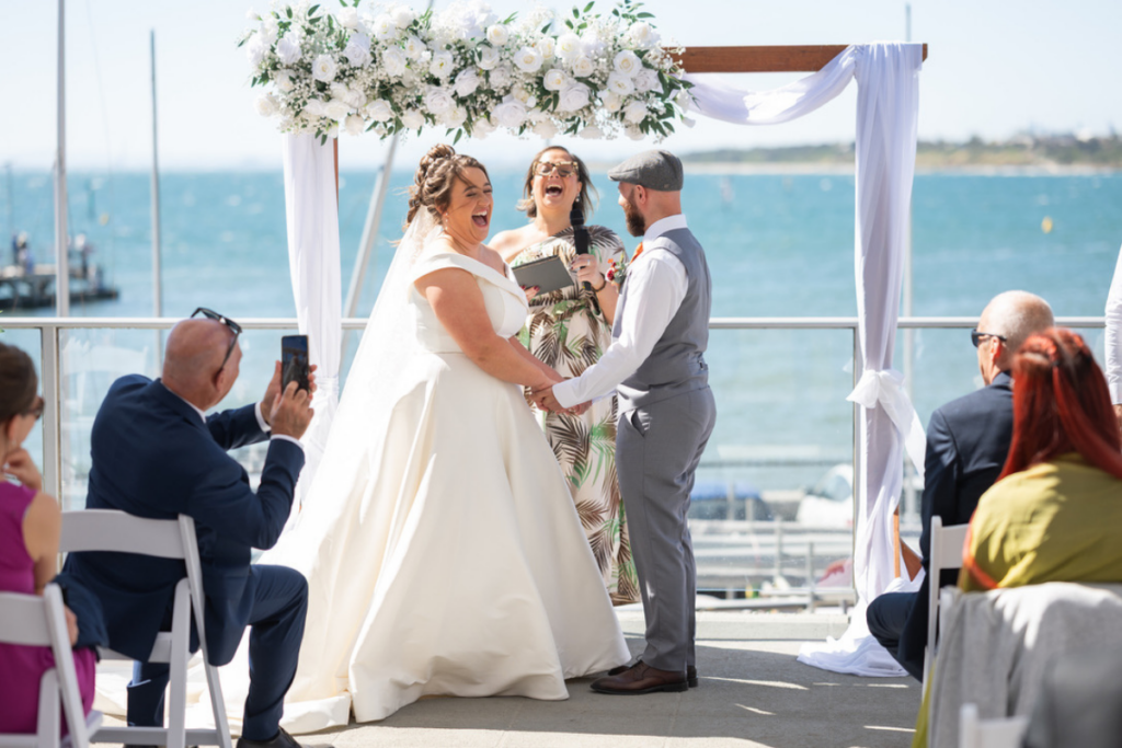 Celebrant Mel Calia laughing with bride and groom during a ceremony by the water